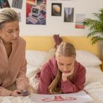 A mother and daughter talk over illustrated materials in a cozy bedroom setting.