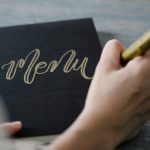 Close-up of a person writing 'Menu' in gold calligraphy on black cardstock with a gold marker.